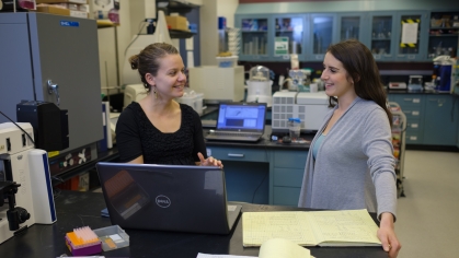 Female professor in a lab talks to a female student. Both are white. The one on the left has her hair in a bun wearing a black top. The student on the right has long brown hair and is wearing a gray sweater.