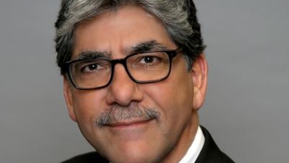 Headshot of man with salt and pepper hair, dark glasses, wearing a black suit, white shirt and green striped tie.