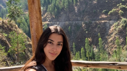 Young woman with long dark hair sitting with a back pack with a view of trees and mountains behind her.