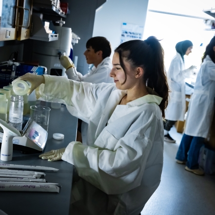 Four students working in a lab all wearing lab coats and doing different tasks.