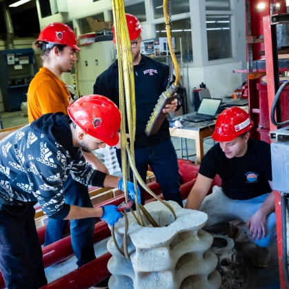 Three male students wearing red hard hats working with an interior jib crane to attach and lift moleded concrete 
