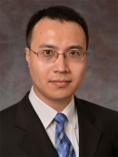 Asian male with eyeglasses wearing a dark suit, white shirt, and a blue patterned tie