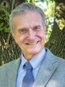 head shot of man with short grey hair wearing a light blue suit, light blue shirt, and blue patterned tie