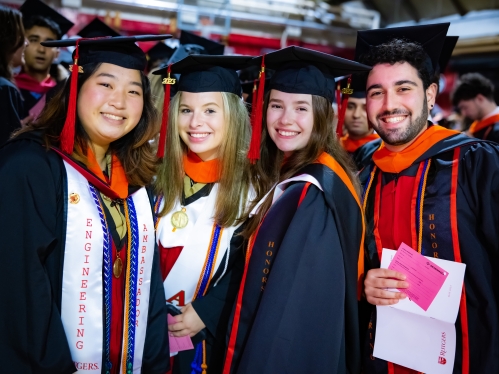 Four students in cap and gowns at graduation ceremony. Three women and a male student.