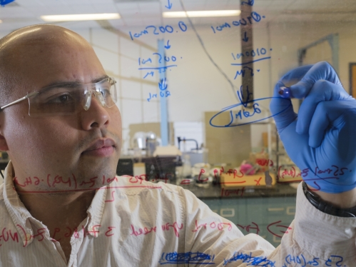 Student wearing safety goggles and blue gloves writes a formula on glass wall.