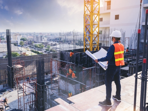 Man in orange and yellow vest with a hard hat stands at a constructions site looking at plans.