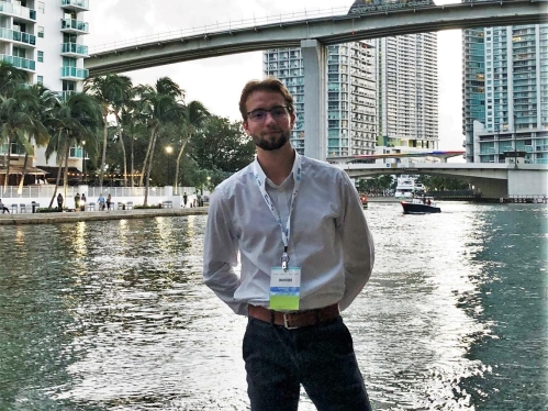 White male college students stands alongside a waterway with a city view behind him. He's wearing dark pants and a white shirt.