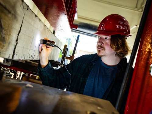 White male graduate student wearing a red hard had with the Rutgers logo holds a device to a beam in a construction lab. He has red hair and is wearing a green and navy blue flannel shirt.