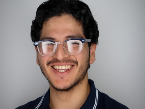 Head shot of a white male college student with black framed glasses, black hair and a beard. He is wearing a navy blue polor shirt.