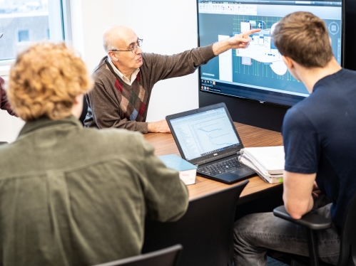 Professor instructing a small grop of three students as he points to the monitor screen,