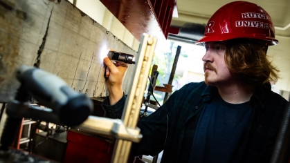White male student in a red Rutgers hard hat holds a flashlight to a concrete beam in a lab setting.
