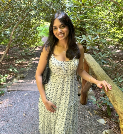 Student poses outdoors with trees in the background. She has long black hair and is wearing a yellow sundress.