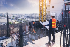 Man in orange and yellow vest with a hard hat stands at a constructions site looking at plans.