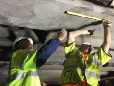 Two men in yellow safety vests and hard hats examine concrete ceiling.